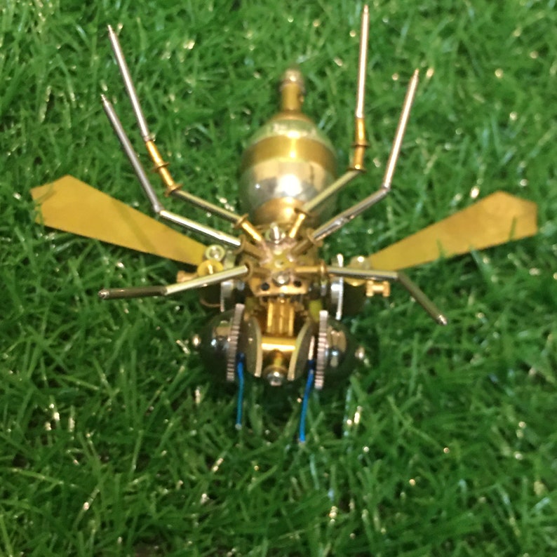 May include: A gold and silver metal insect sculpture with wings and long legs. The insect is made of various gears and other mechanical parts.