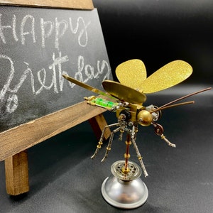 May include: A steampunk-style insect sculpture with gold and green accents. The insect has wings made of gold glitter paper and is standing on a silver base. The sculpture is in front of a small chalkboard with the words "Happy Birthday" written on it.