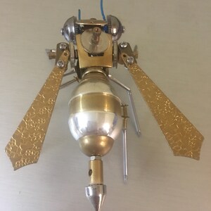 May include: A gold and silver steampunk-style insect sculpture with intricate details. The insect has a pointed tail, two wings, and a gear-like head.