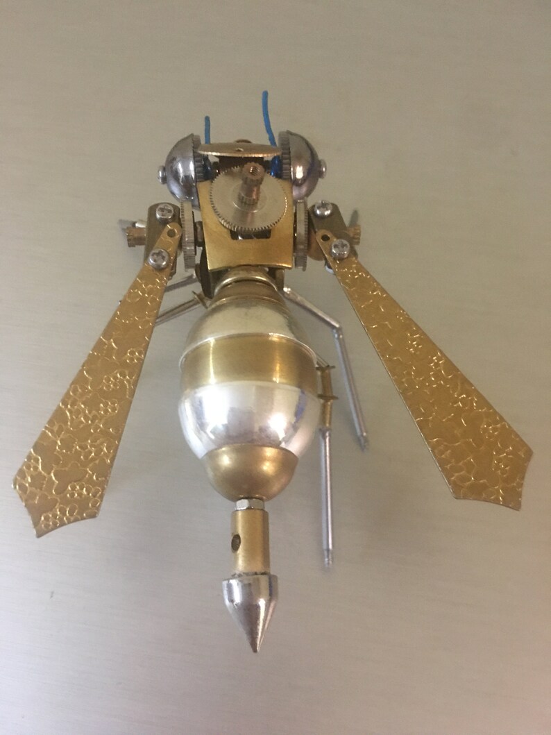 May include: A gold and silver steampunk-style insect sculpture with intricate details. The insect has a pointed tail, two wings, and a gear-like head.