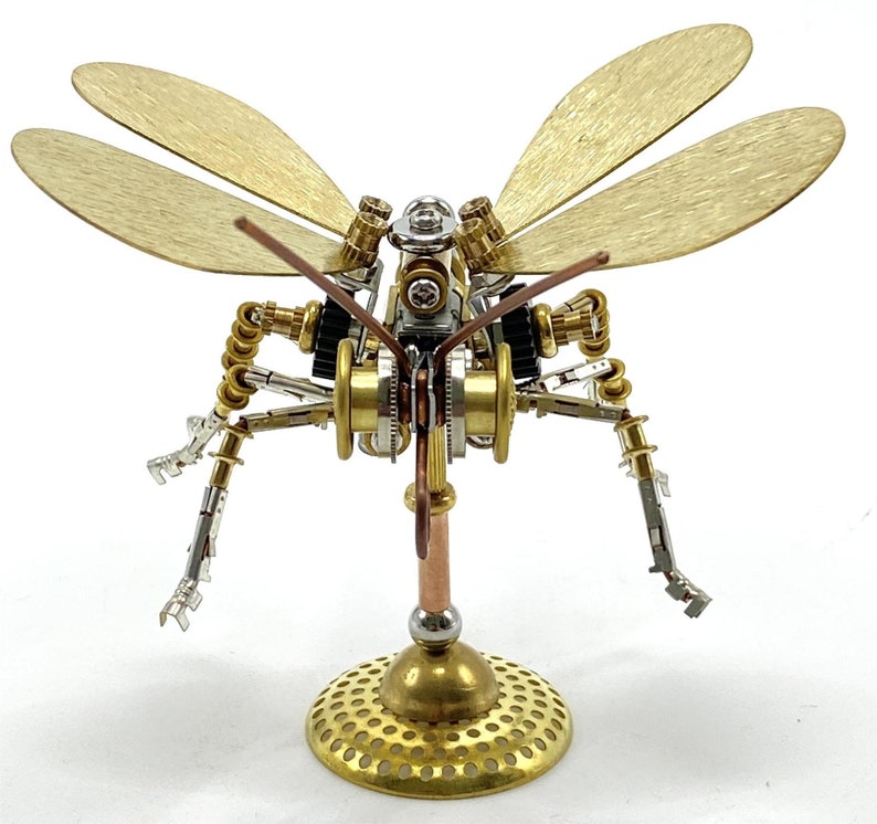 May include: A steampunk dragonfly sculpture crafted from brass, copper, and silver-toned metal. The wings are a brushed gold colour, and the body features intricate gears and mechanical details. The dragonfly is mounted on a round, perforated brass base.