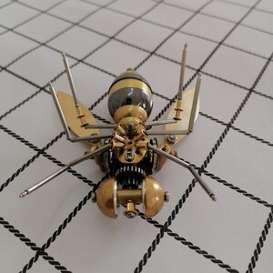 May include: A detailed, intricate, steampunk-style metal wasp sculpture with a gold and silver finish. The wasp is posed with its wings spread and its legs extended.