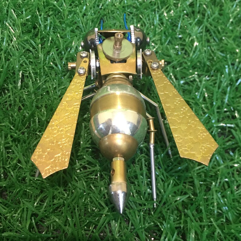 May include: A gold and silver steampunk-style wasp figurine with intricate details and a pointed tail. The figurine is set against a green grass background.