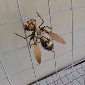 May include: A gold and black metal sculpture of a wasp with intricate details. The sculpture is mounted on a white wire grid.