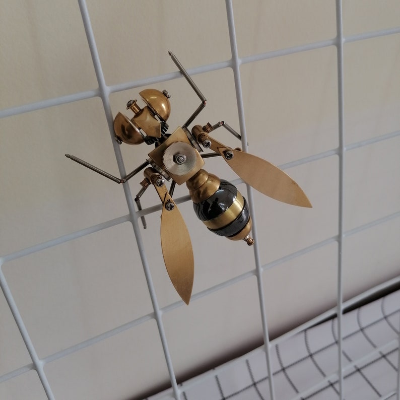 May include: A gold and black metal sculpture of a wasp with intricate details. The sculpture is mounted on a white wire grid.