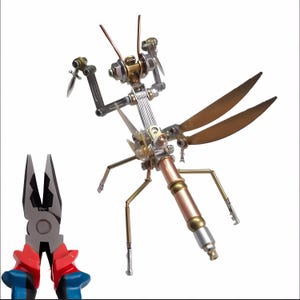 May include: A metal sculpture of a praying mantis with wings, made from various metal parts, including copper, brass, and silver. The sculpture is standing on a white surface. A pair of red and blue pliers are in the foreground.