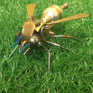 May include: A detailed, steampunk-style metal sculpture of a wasp with gold and silver accents. The sculpture is posed on a green grassy surface.
