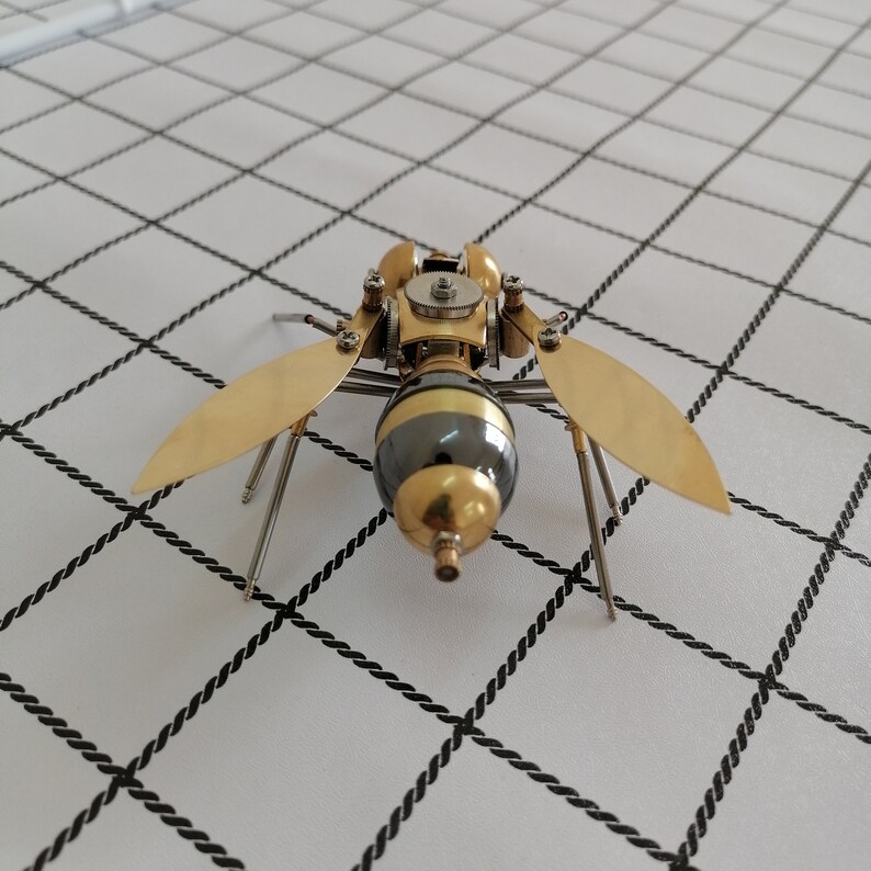 May include: A gold and black metal insect sculpture with intricate details. The insect has six legs and two wings. The wings are made of thin metal and are shaped like a wasp's wings.