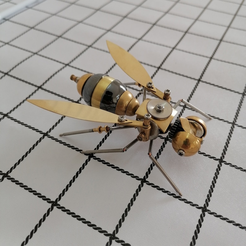 May include: A detailed, steampunk-style wasp made of metal and gears. The wasp is gold and black with intricate details and is posed on a white surface with black grid lines.