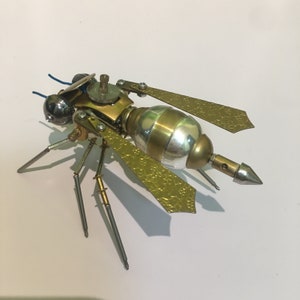 May include: A gold and silver steampunk-style insect sculpture with intricate details. The insect has six legs, two wings, and a long, pointed tail.