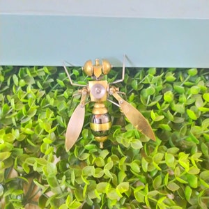 May include: A gold and silver steampunk-style bee sculpture with intricate details. The bee is perched on a bed of green artificial foliage.