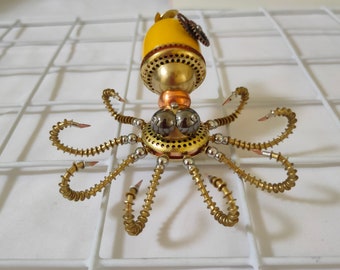 May include: A steampunk-style octopus sculpture made of metal with a yellow head and eight articulated arms. The sculpture has a detailed design with gears and other mechanical elements.