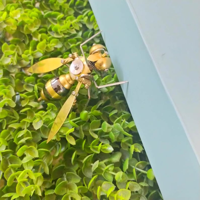 May include: A gold and silver steampunk-style wasp sculpture with intricate details. The wasp is perched on a green artificial grass surface.