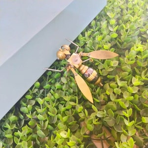 May include: A gold and black metal insect sculpture with intricate details. The sculpture is made to look like a wasp or bee and is sitting on a bed of green artificial grass.