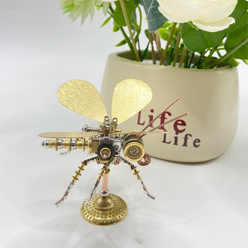 May include: A detailed steampunk dragonfly sculpture crafted from brass, copper, and silver-toned metal. The insect features intricate mechanical elements, including gears and textured wings. The sculpture is mounted on a circular base and is a decorative item.