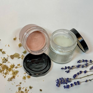 May include: Two small glass jars with black lids containing a light pink and a light green cream. The jars are on a white surface with dried flowers and lavender sprigs.