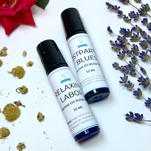 May include: Two essential oil roller bottles with black caps and white labels. One reads "RELAXIN LABOR" and the other "POSTPARTUM BLUES." Both are 10 ml. Dried lavender, chamomile, and a red rose are in the background.