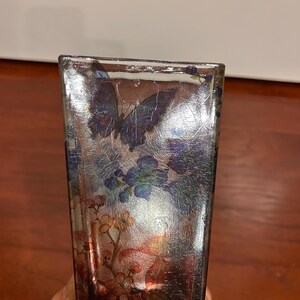 Pu&ograve; includere: Un vaso rettangolare in vetro con finitura argentata screpolata. Il vaso &egrave; decorato con immagini di farfalle e motivi floreali nei toni del blu, viola e arancione. Il vaso &egrave; un oggetto decorativo.