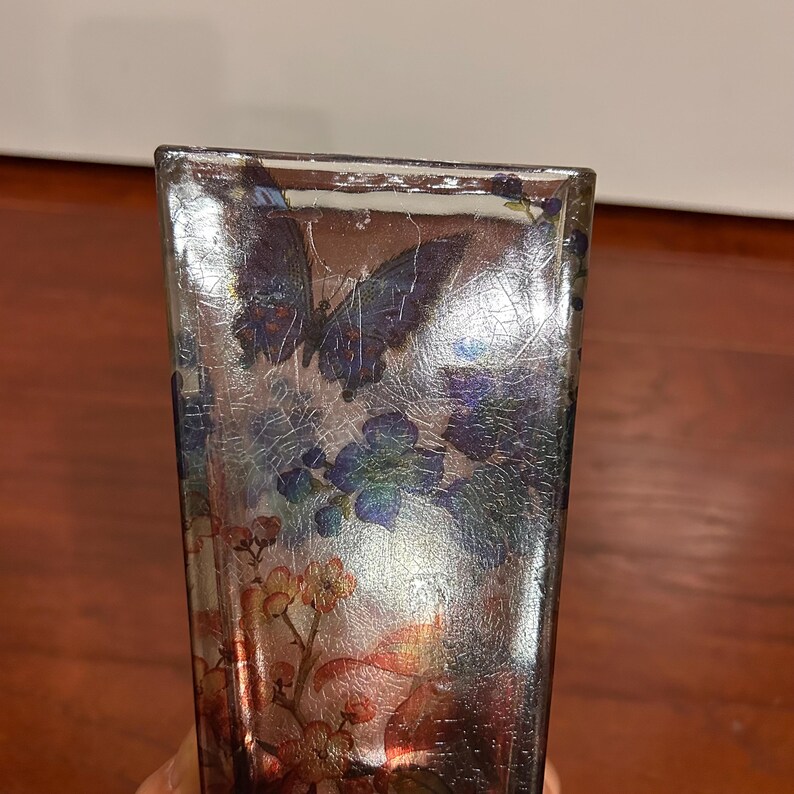 Pu&ograve; includere: Un vaso rettangolare in vetro con finitura argentata screpolata. Il vaso &egrave; decorato con immagini di farfalle e motivi floreali nei toni del blu, viola e arancione. Il vaso &egrave; un oggetto decorativo.