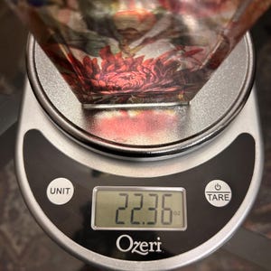 Pu&ograve; includere: Una ciotola decorativa in vetro con un motivo floreale, appoggiata su una bilancia da cucina digitale. La bilancia mostra un peso di 634.05 grammi. La bilancia &egrave; argentata e nera, con il marchio "Ozeri" visibile.