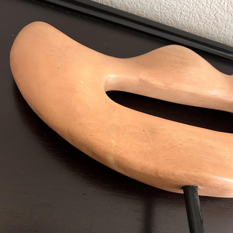 May include: A light peach-colored, abstract wooden sculpture with a large, oval-shaped opening. The sculpture is mounted on a black metal stand. The wood grain is visible on the surface. The artwork is a decorative home accent.