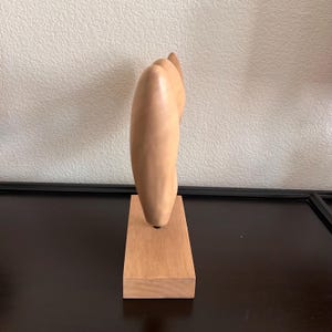 May include: A light-colored wooden sculpture of a hand with fingers pointing upwards. The sculpture is mounted on a rectangular wooden base. The artwork is displayed on a dark surface against a neutral wall.