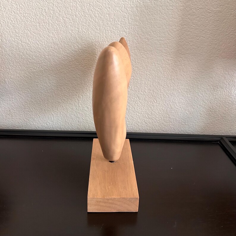 May include: A light-colored wooden sculpture of a hand with fingers pointing upwards. The sculpture is mounted on a rectangular wooden base. The artwork is displayed on a dark surface against a neutral wall.