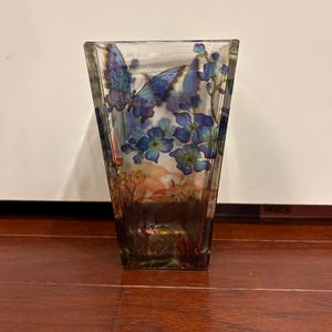 Pu&ograve; includere: Un vaso di vetro trasparente a forma trapezoidale. Il vaso &egrave; decorato con farfalle blu e fiori blu. Il fondo del vaso presenta un motivo floreale marrone e arancione. Il vaso &egrave; appoggiato su una superficie di legno.