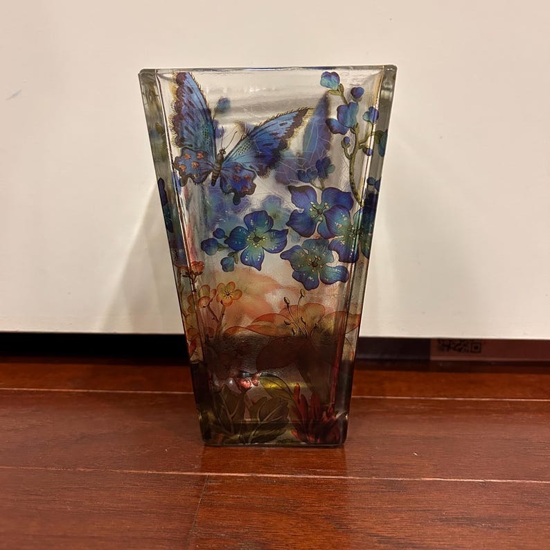 Pu&ograve; includere: Un vaso di vetro trasparente a forma trapezoidale. Il vaso &egrave; decorato con farfalle blu e fiori blu. Il fondo del vaso presenta un motivo floreale marrone e arancione. Il vaso &egrave; appoggiato su una superficie di legno.