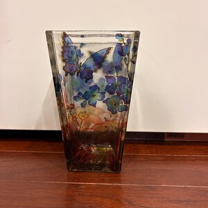 Pu&ograve; includere: Un vaso trasparente quadrato con la parte superiore svasata, adornato con un design a farfalla e floreale. La farfalla &egrave; principalmente blu con dettagli intricati sulle ali. I fiori sono blu e il design ha un effetto acquerello.