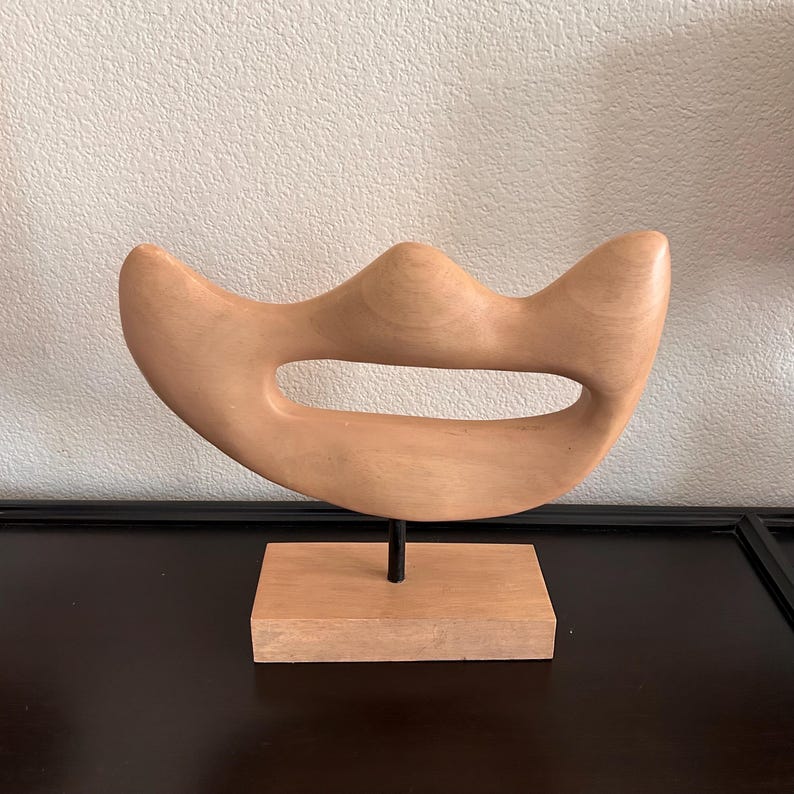 May include: A light-colored wooden sculpture with a curved, abstract design. The sculpture has a central opening and is mounted on a small, rectangular wooden base with a black metal rod. The piece is displayed on a dark surface.
