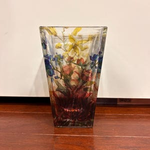 Pu&ograve; includere: Un vaso di vetro trasparente e quadrato con un motivo floreale. Il vaso presenta una colorata composizione di fiori nelle tonalit&agrave; del rosso, giallo, blu e verde. I fiori sono dipinti all'interno del vetro, creando un effetto a strati. Il vaso &egrave; appoggiato su una superficie di legno.