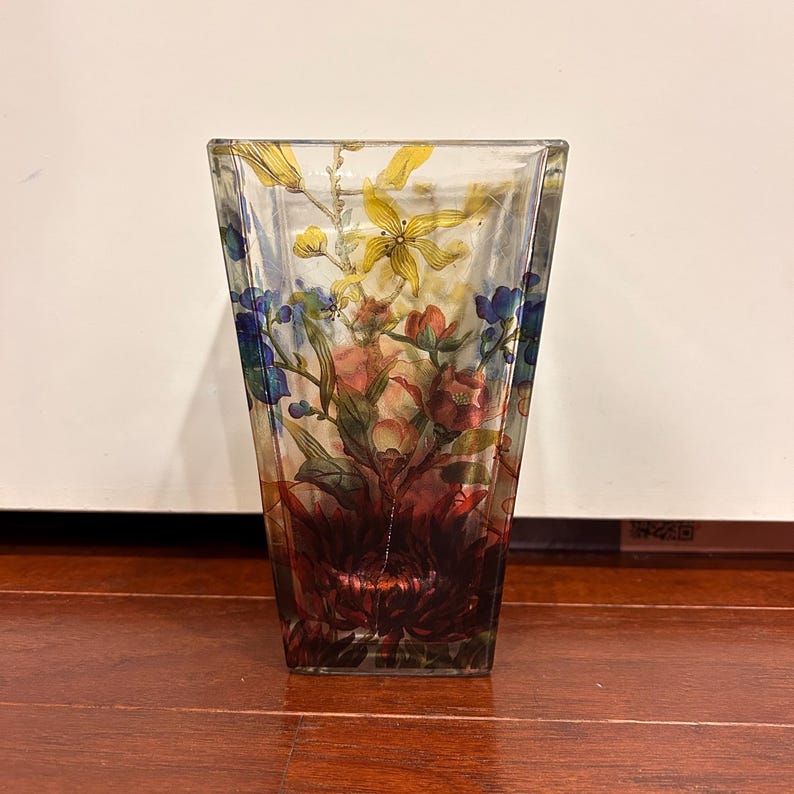 Pu&ograve; includere: Un vaso di vetro trasparente e quadrato con un motivo floreale. Il vaso presenta una colorata composizione di fiori nelle tonalit&agrave; del rosso, giallo, blu e verde. I fiori sono dipinti all'interno del vetro, creando un effetto a strati. Il vaso &egrave; appoggiato su una superficie di legno.