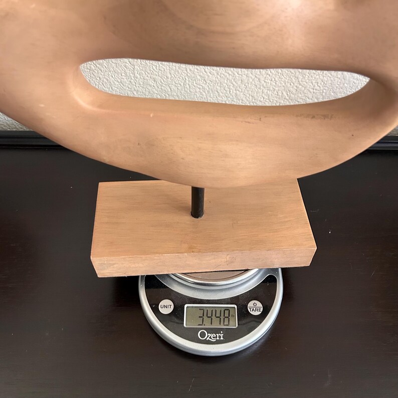 May include: A light brown wooden sculpture with a curved, open design. The sculpture is mounted on a small wooden base with a metal rod. The base rests on a digital scale displaying a weight of 3.448.