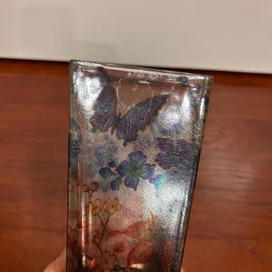 Pu&ograve; includere: Un vaso rettangolare in vetro con finitura argentata e un design a farfalle e fiori. Il vaso presenta una texture screpolata e accenti iridescenti, con elementi floreali blu, rossi e gialli.