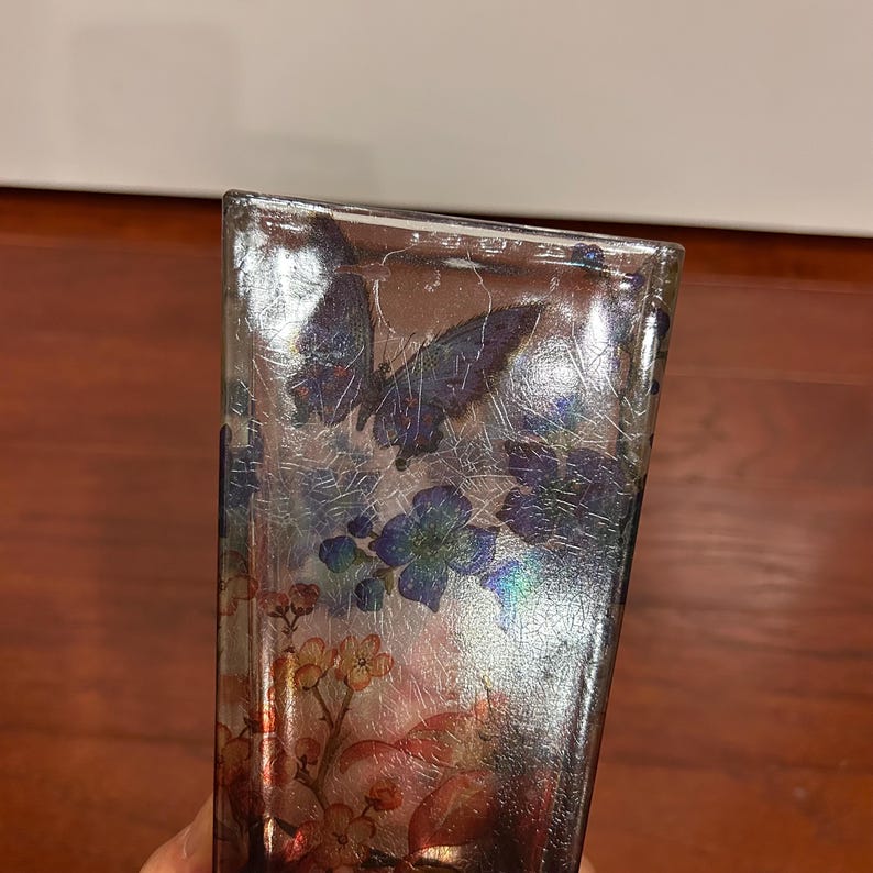 Pu&ograve; includere: Un vaso rettangolare in vetro con finitura argentata e un design a farfalle e fiori. Il vaso presenta una texture screpolata e accenti iridescenti, con elementi floreali blu, rossi e gialli.