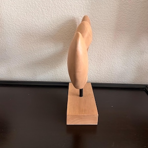 May include: A light-colored wooden sculpture with a smooth, curved design. The sculpture is mounted on a rectangular wooden base with a black metal rod. The artwork is displayed against a neutral background.