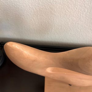 May include: A light-colored wooden shoe last, showcasing a smooth, curved design. The last is positioned on a wooden base, with a natural wood grain visible. The object is designed for shoemaking.