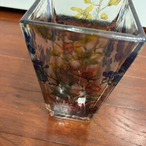 Pu&ograve; includere: Un vaso di vetro trasparente di forma quadrata con un disegno floreale. I fiori sono nelle tonalit&agrave; del rosso, del rosa, del giallo e del blu. Il vaso &egrave; appoggiato su una superficie di legno.
