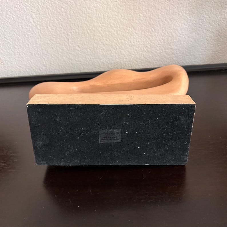 May include: A light brown, abstract sculpture with a smooth, curved design. The sculpture sits atop a rectangular base with a black, felt-like surface. The base is made of a light-colored wood.