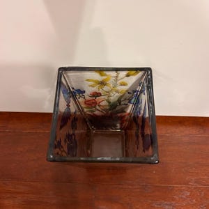 Pu&ograve; includere: Un vaso quadrato di vetro con un motivo floreale. Il vaso ha un corpo trasparente con un bordo scuro. Il motivo floreale presenta fiori gialli, rossi e blu. Il vaso &egrave; appoggiato su una superficie di legno.