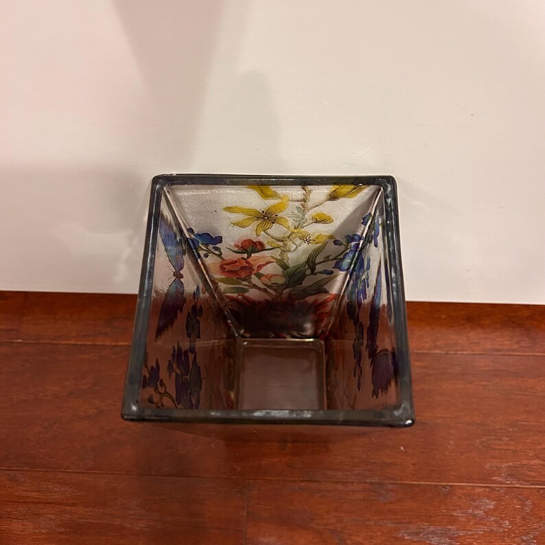 Pu&ograve; includere: Un vaso quadrato di vetro con un motivo floreale. Il vaso ha un corpo trasparente con un bordo scuro. Il motivo floreale presenta fiori gialli, rossi e blu. Il vaso &egrave; appoggiato su una superficie di legno.