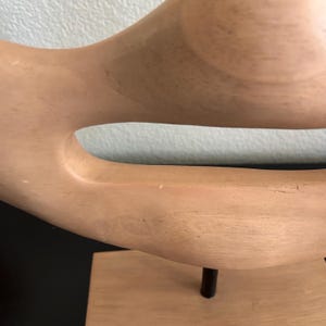 May include: A light-colored wooden sculpture with a curved, organic shape. The sculpture has a smooth finish and is mounted on a rectangular base with a dark metal rod. The design features a large opening in the center.