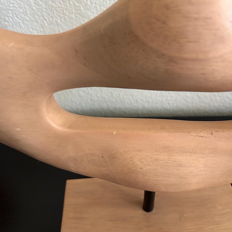 May include: A light-colored wooden sculpture with a curved, organic shape. The sculpture has a smooth finish and is mounted on a rectangular base with a dark metal rod. The design features a large opening in the center.