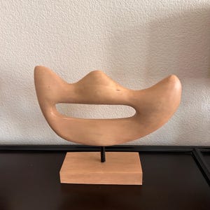 May include: A light brown, abstract wooden sculpture with a curved shape and a central oval opening. The sculpture is mounted on a small, rectangular wooden base with a black metal rod. The artwork is displayed against a neutral background.