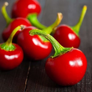 May include: A group of red, cherry-shaped peppers with green stems on a dark wooden surface.