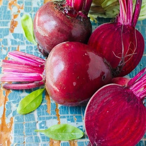 May include: Four red beetroot with green leafy tops on a blue and brown textured surface. The beetroot are whole and have their stems attached.