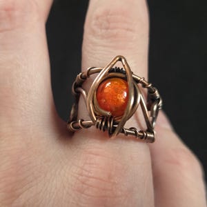 Puede incluir: Un anillo de alambre de color cobre oscuro, con una cuenta redonda naranja en el centro que presenta una superficie texturizada y moteada. El alambre de metal está enrollado y formado en un marco geométrico alrededor de la piedra.