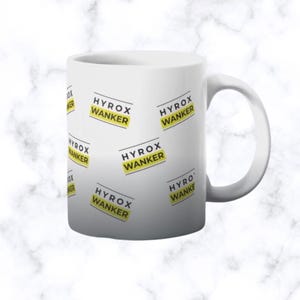 May include: White ceramic mug featuring the text "HYROX WANKER" in black and yellow, repeated across the surface. The mug has a white handle. The background is a marble pattern.