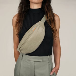 Könnte beinhalten: Eine hellgrüne Cord-Bauchtasche mit einem braunen Gurt. Die Bauchtasche wird von einer Person getragen, die ein schwarzes Shirt mit Stehkragen und eine grüne Hose trägt.